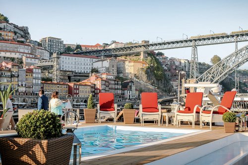 Sonnendeck der A-ROSA ALVA mit Pool, weiß-roten Liegestühlen und Pflanzen, im Hintergrund eine Brücke in Porto und die bunten Häuser der Stadt.