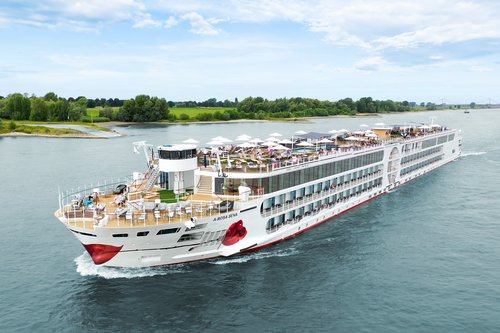 Die A-ROSA SENA fährt über den Rhein, das mit Liegestühlen und Sonnenschirmen ist gut besucht im Hintergrund das Ufer mit grüner Landschaft und Bäumen.
