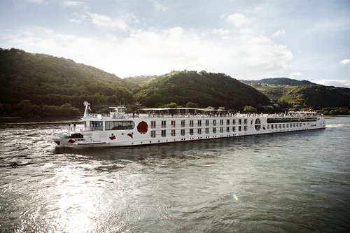 Die A-ROSA BRAVA gleitet am Nachmittag auf dem Rhein entlang, flankiert von grünen Hügeln und kleinen Siedlungen im Hintergrund.