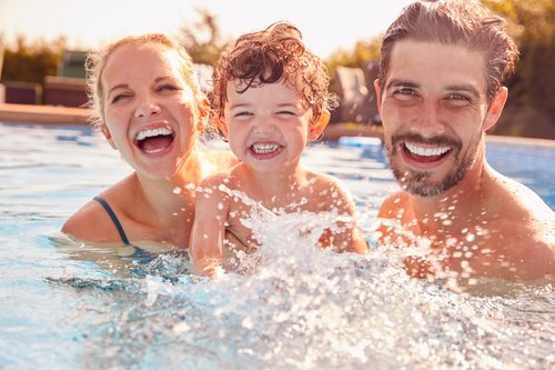 Lachende Familie mit Mutter, Vater und kleinem Kind plantscht gemeinsam im Schwimmbecken.