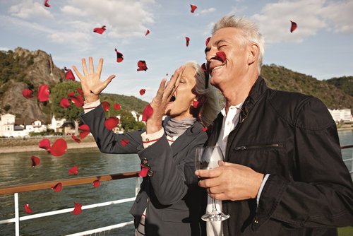 Ein älteres Paar steht auf dem Sonnendeck eines Flusskreuzfahrtschiffs. Die Frau wirft Rosenblüten in die Luft und der Mann trinkt ein Glas Wein. Beide freuen sich. Im Hintergrund die Hügellandschaft der Loreley am Rhein.