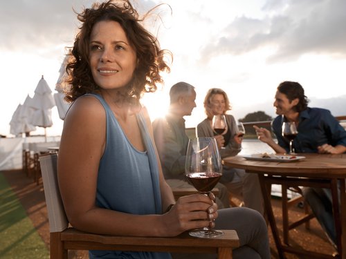 Vier Personen sitzen auf dem Deck eines Flusskreuzfahrtschiffs und genießen einen Rotwein bei einem Gespräch bei Sonnenschein. Die Frau im Vordergrund sitzt abgewandt und schaut in die Ferne.