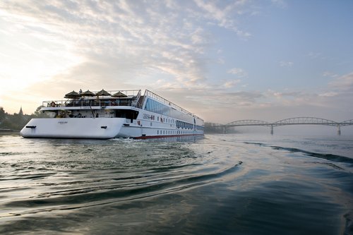 Heckansicht des Flusskreuzfahrtschiffs A-ROSA MIA auf der Donau bei Sonnenaufgang. Im Vordergrund ist der Fluss mit leichten Wellen. Im Hintergrund liegt eine Brücke.
