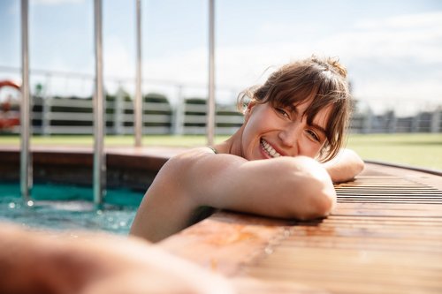 Eine Frau im Pool auf dem Deck eines Flusskreuzfahrtschiffes lehnt entspannt die Arme und den Kopf auf den Poolrand und lächelt die Person neben sich an.