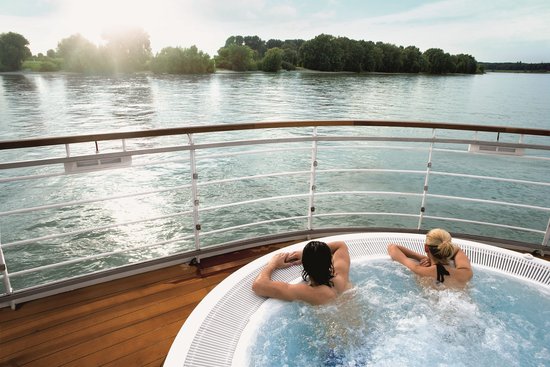 Zwei Personen entspannen in einem Whirlpool auf dem Deck eines Flusskreuzfahrtschiffs mit Ausblick auf den Rhein bei Sonnenschein.