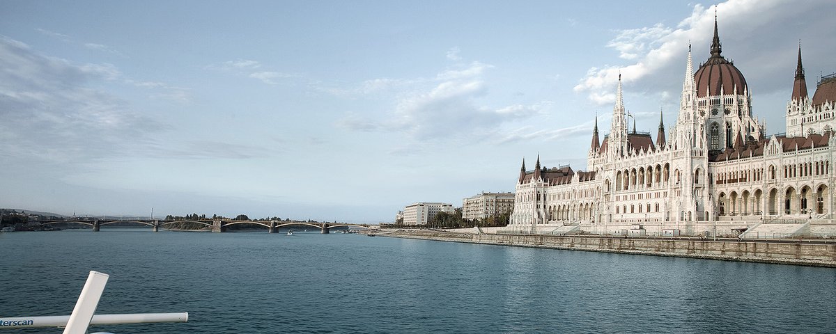 Blick von einem Kreuzfahrtschiff auf das Parlamentsgebäude in Budapest am Donauufer unter bewölktem Himmel.