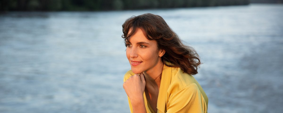 Eine elegant gekleidete Frau lehnt an der Reling eines Flusskreuzfahrtschiffes und betrachtet den Sonnenuntergang über dem Fluss, während sie entspannt die Aussicht genießt.