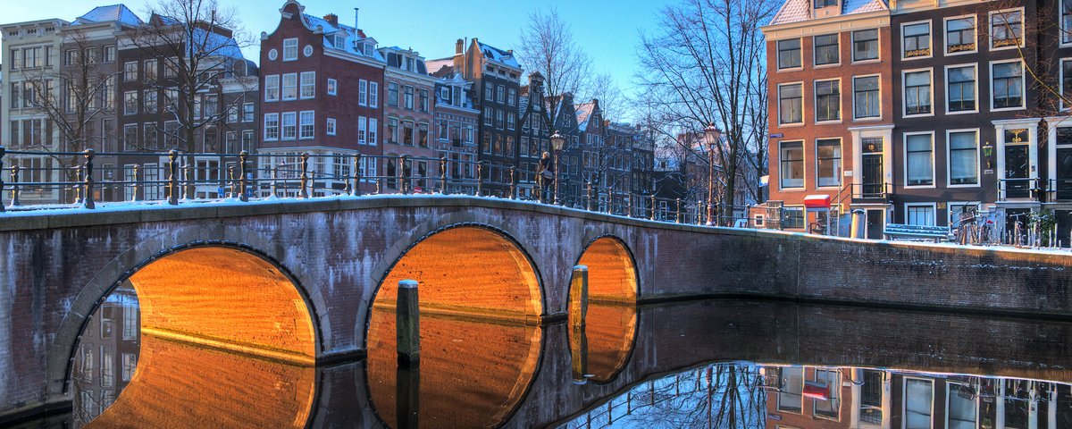 Brücke mit drei Bögen über einen Kanal in Amsterdam, beleuchtet von warmem Licht, mit historischen Häusern und klarer Spiegelung im Wasser
