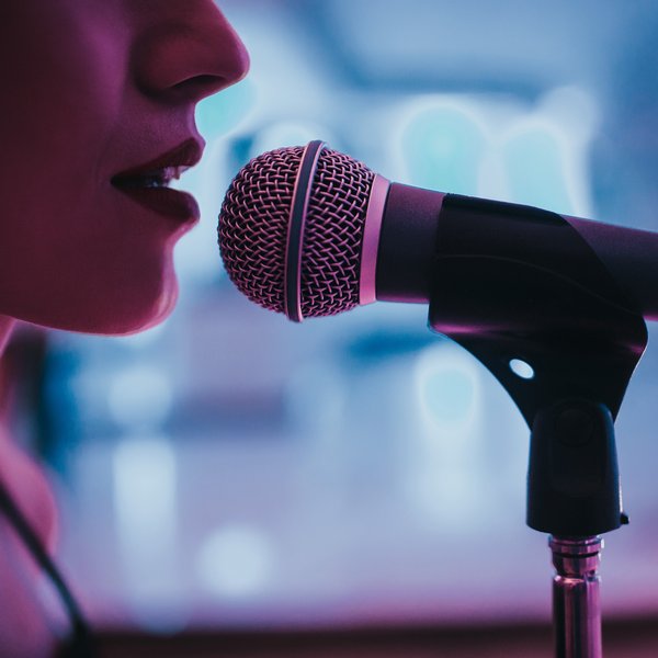 Nahaufnahme einer Sängerin, die in ein Mikrofon singt, mit unscharfem Hintergrund und blauer Beleuchtung.