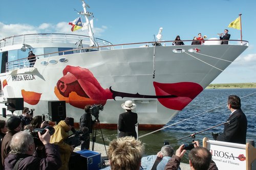 Bug des Flusskreuzfahrtschiffs A-ROSA BELLA mit großem Kussmund und einer Rose als Schiffsbemalung bei der Taufe, Fotografen und Zuschauer im Vordergrund.