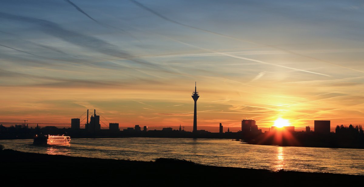 Blick auf die Düsseldorfer Skyline bei Sonnenuntergang, mit dem Rheinturm und modernen Gebäuden am Flussufer.