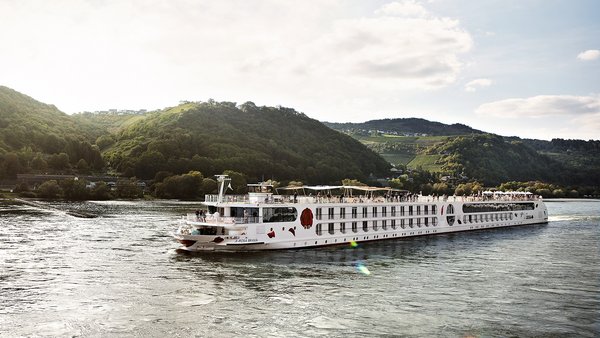 Die A-ROSA BRAVA gleitet am Nachmittag auf dem Rhein entlang, am Ufer grüne, bewaldete Hügeln und kleinen Siedlungen im Hintergrund.