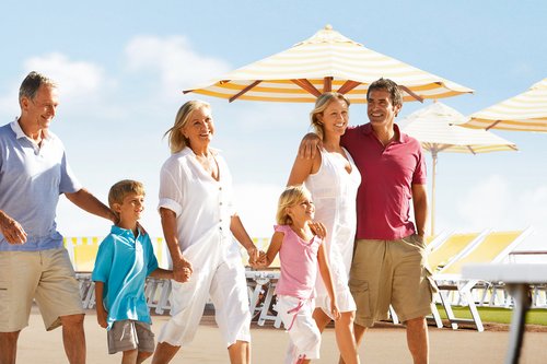 Drei Generationen Familie spazieren lachend auf einem Sonnendeck mit gelb-weißen Sonnenschirmen und Liegestühlen.