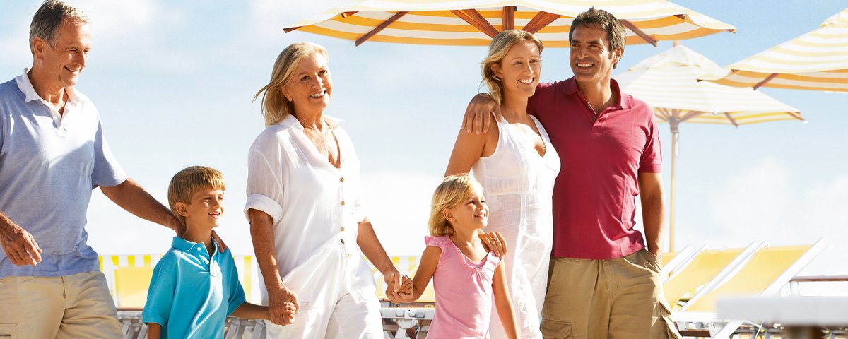 Drei Generationen Familie spazieren lachend auf einem Sonnendeck mit gelb-weißen Sonnenschirmen und Liegestühlen.
