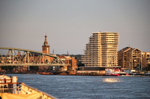 Blick auf die Uferpromenade von Nimwegen mit einem modernen hohen Wohngebäude, einer historischen Kirchturmspitze und einer Brücke über den Fluss.