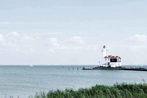 Ein Leuchtturmhaus am Ijsselmeer mit Schilf im Vordergrund und Segelbooten auf dem Meer, unter einem bewölkten Himmel.