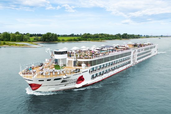 Die A-ROSA SENA fährt über den Rhein, das mit Liegestühlen und Sonnenschirmen ist gut besucht im Hintergrund das Ufer mit grüner Landschaft und Bäumen. 