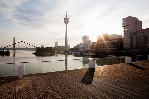 Blick von einer Holzbrücke mit Glasgeländer auf Düsseldorf mit dem Rheinturm und modernen Gebäuden am Flussufer, bei Sonnenschein und dem reflektierenden Rhein mit Brücken im Hintergrund.