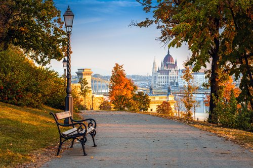 Herbstliche Ansicht des Parlamentsgebäude von Budapest im Hintergrund, gesehen von einem laubbedeckten Park. Im Vordergrund steht eine einzelne Parkbank.