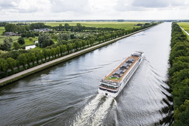 Ein Flusskreuzfahrtenschiff fährt auf dem Rhein am Ufer ein kleines Dorf, grüne Felder und Bäume. Das Sonnendeck ist mit Whirlpool, Sonnenliegen und Sonnensegeln ausgestattet.