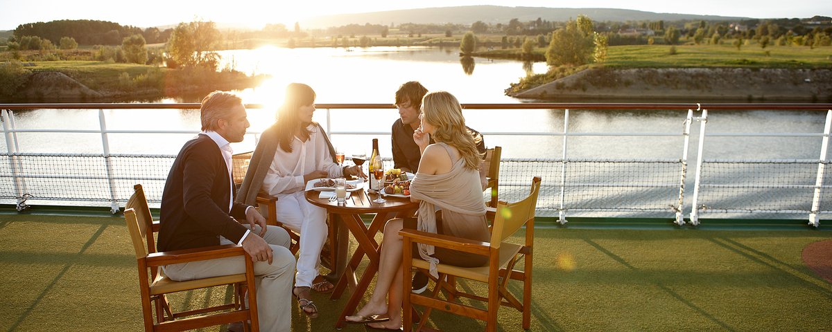 Zwei Paare sitzen an einem Tisch auf dem Sonnendeck, genießen Wein und etwas zu essen bei Sonnenuntergang, umgeben von grünen Uferlandschaften und einer warmen Abendstimmung.
