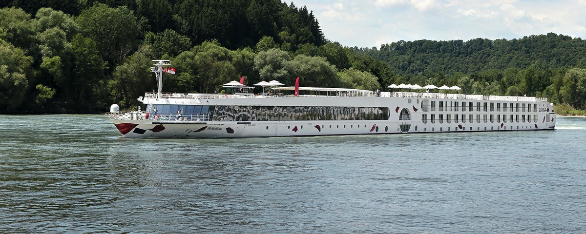 Die A-ROSA SILVA fährt am Nachmittag auf dem Rhein entlang am Ufer grüne, bewaldete Hügeln, der Himmel ist blau und leicht bewölkt.