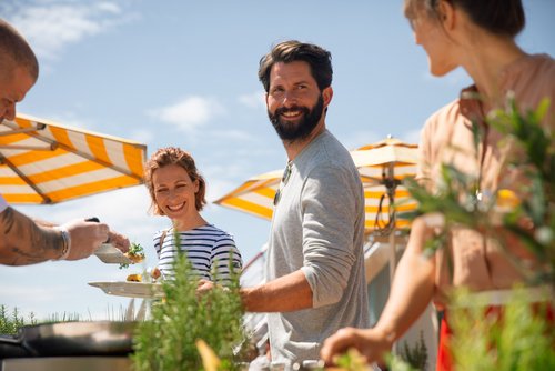 Gruppe von Menschen bei einem BBQ im Freien, eine Frau hält einen Teller, ein Mann lächelt in die Kamera, gelb-weiße Sonnenschirme im Hintergrund.