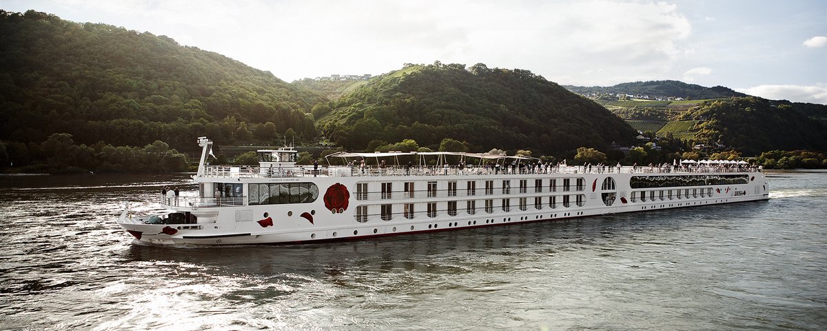 Ein Schiff der A-ROSA Rheinflotte gleitet am Nachmittag auf dem Rhein entlang, am Ufer grüne, bewaldete Hügeln und kleinen Siedlungen im Hintergrund.