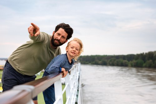 Vater und Sohn stehen auf dem Deck eines Flusskreuzfahrtschiffs und blicken über die Reling auf den Fluss. Der Vater zeigt auf etwas in der Ferne.