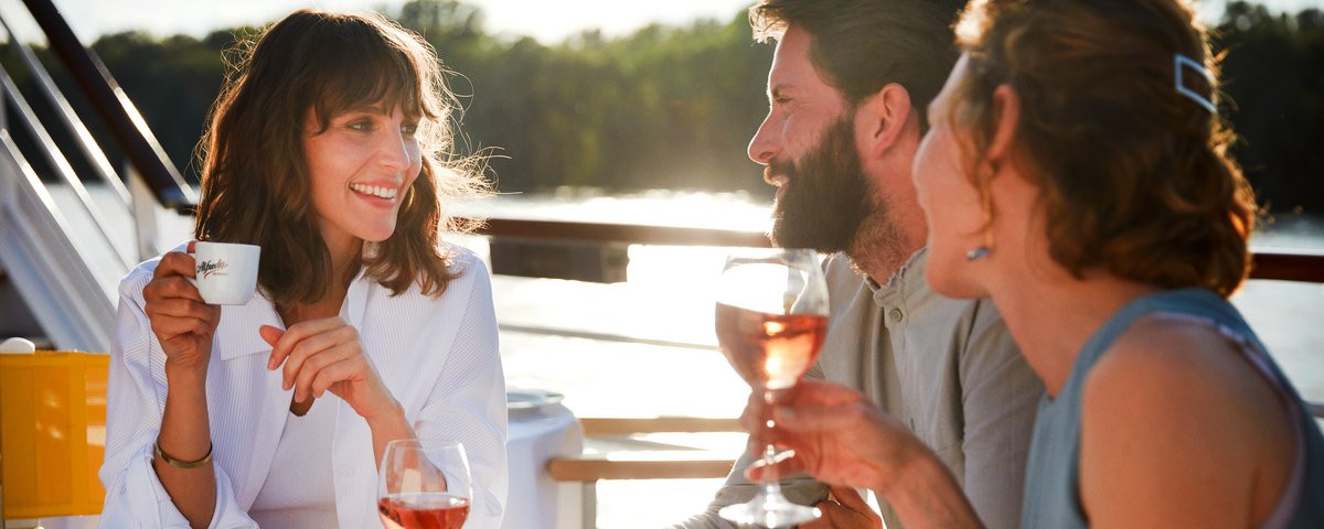 Drei Personen sitzen an einem gedeckten Tisch im Freien, trinken Wein und unterhalten sich bei Sonnenlicht.