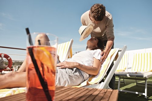 Eine Frau liegt auf einer Sonnenliege auf dem Deck eines Flusskreuzfahrtschiffs. Ein Mann steht hinter ihr und spendet ihr Schatten mit einem Hut. Im Vordergrund steht ein Getränk mit Orangenscheibe.