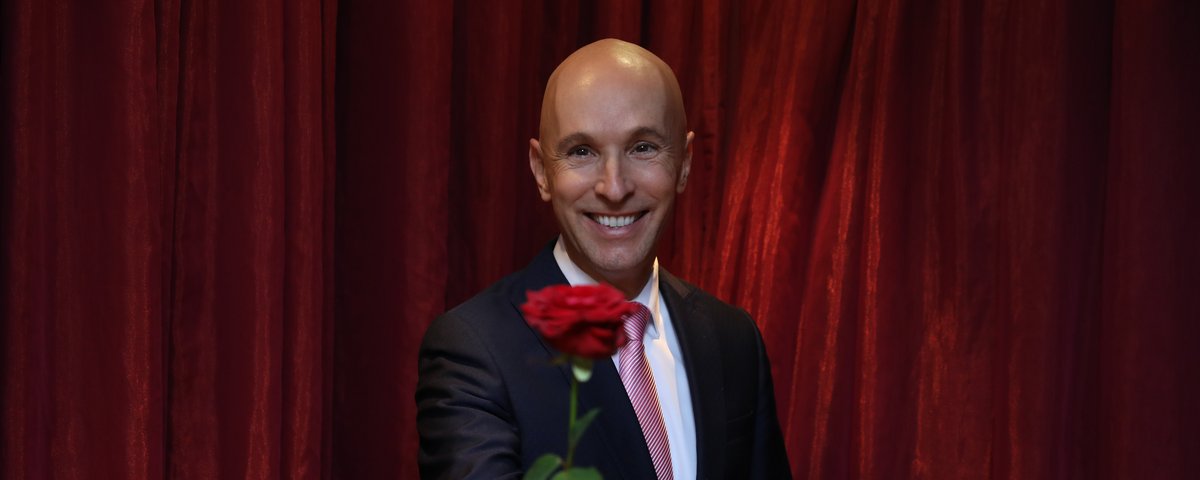 Michael Begasse steht lächelnd in einem eleganten Anzug vor einem Vorgang und hält eine rote Rose in der Hand.