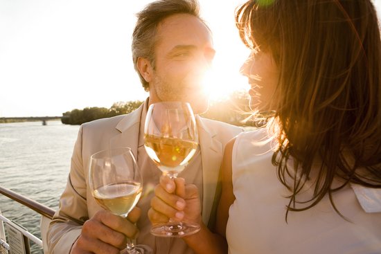 Ein Paar steht bei Sonnenuntergang mit einem Glas Wein in der Hand an der Rehling eines Flussschiffes. Im Hintergrund der Fluss und eine Uferlandschaft. 
