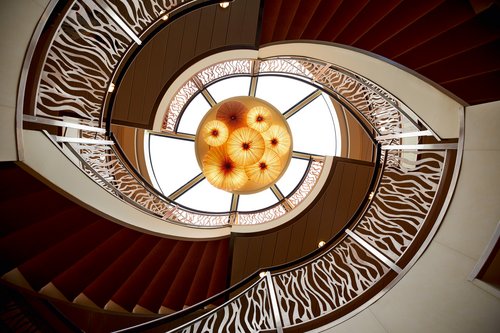 Spiralförmige Treppe mit dekorativem Geländer und runden Lampenschirmen im Atrium eines Schiffs.