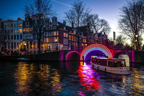 Beleuchtete Brücke mit buntem Neon-Regenbogen über einem Kanal in Amsterdam bei Dämmerung, Boot auf dem Wasser