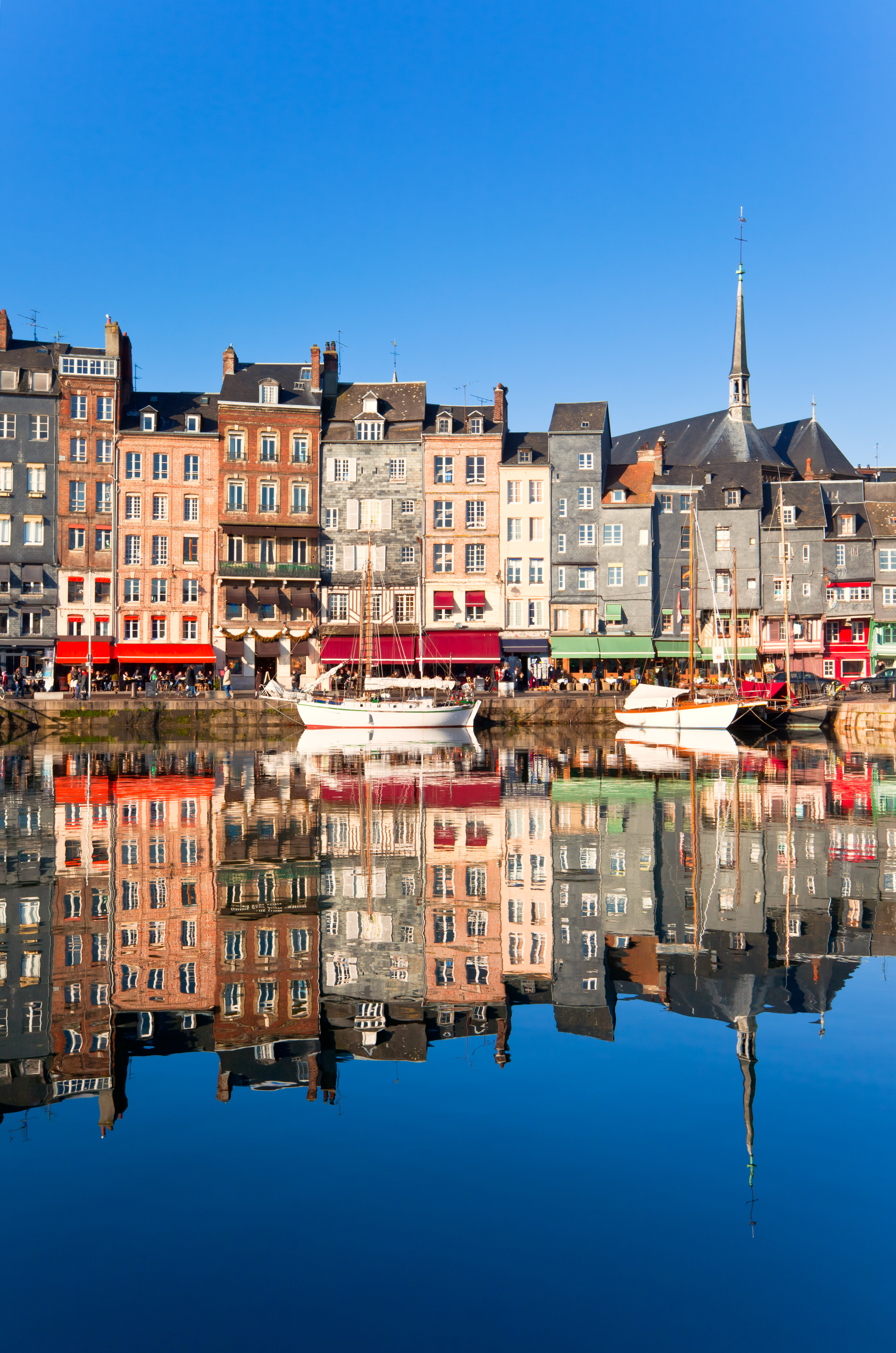 Hafen von Honfleur mit bunten historischen Gebäuden und Segelbooten im Vordergrund, unter einem klaren blauen Himmel.
