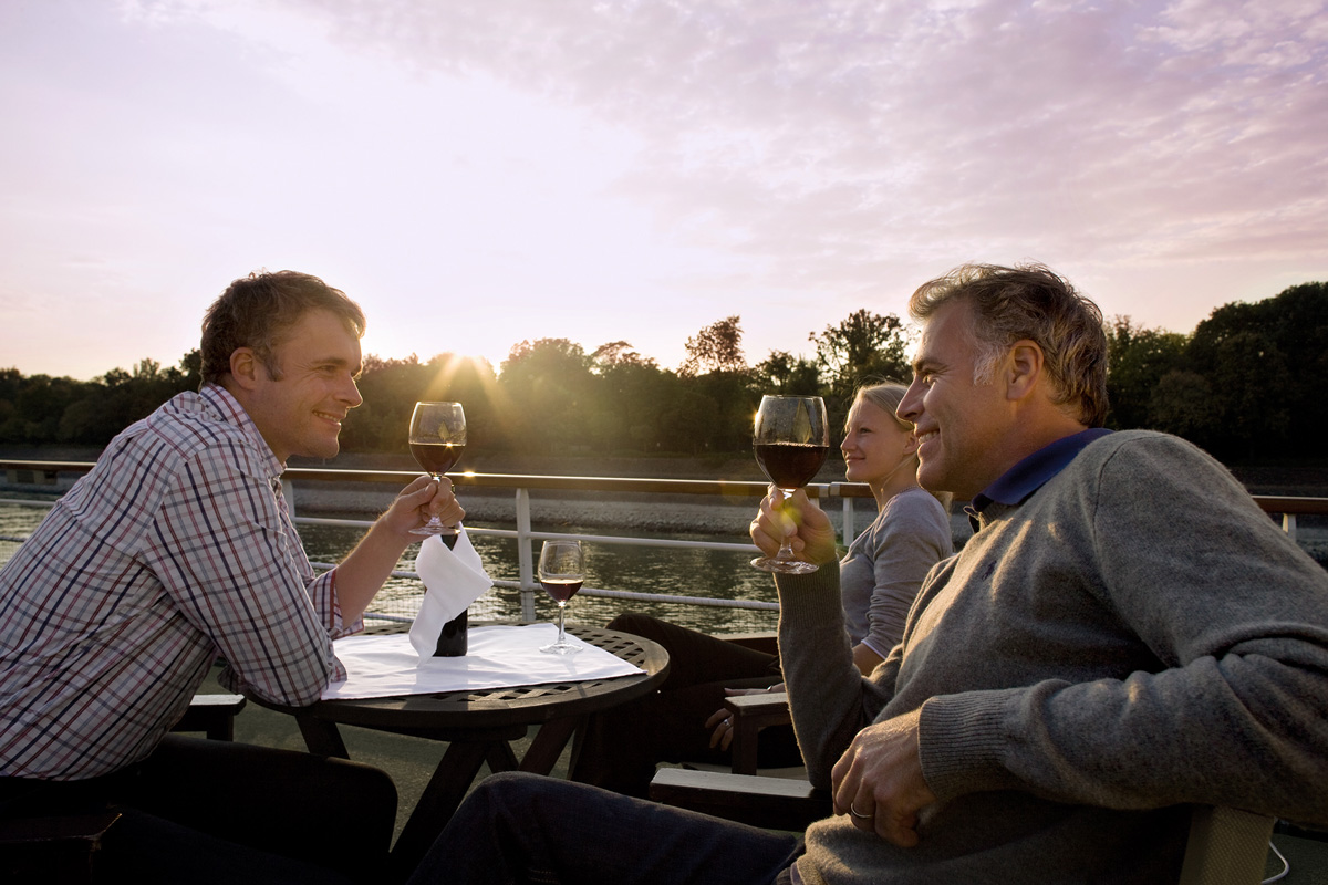 Drei Erwachsene stoßen in der Abendsonne mit Rotweingläsern auf dem Sonnendeck eines A-ROSA Schiffs an.