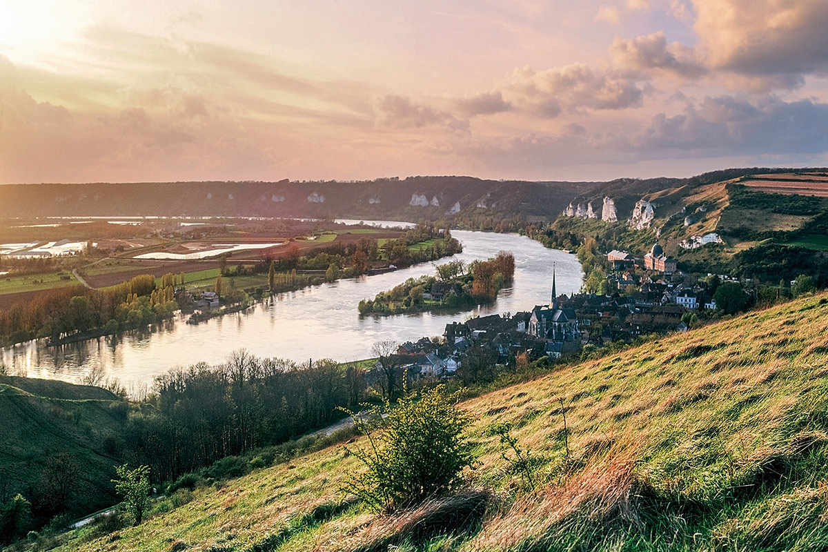Weite Flusslandschaft an der Seine in der goldenen Abendsonne mit hügeliger Umgebung und kleiner Ortschaft am Ufer.